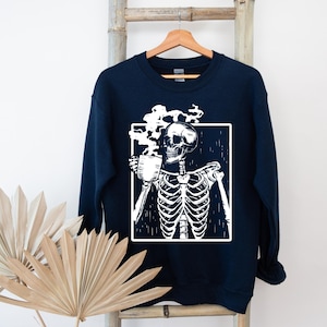 May include: Navy blue sweatshirt with a white graphic of a skeleton enjoying a coffee. The skeleton is inside a white square. The sweatshirt is hanging on a wooden rack. The design is a humorous take on a coffee break.