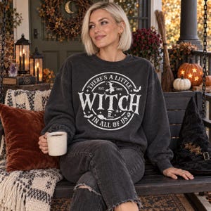 May include: A charcoal grey long-sleeve sweatshirt featuring a white circular graphic with the text "THERE'S A LITTLE WITCH IN ALL OF US" and "EST. 1692," accompanied by illustrations of a witch, a broom, bats, and a skull, worn with dark grey jeans.