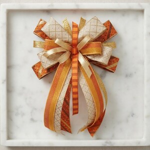May include: A decorative bow crafted from multiple ribbons in shades of orange, gold, and cream. The ribbons feature various textures and patterns, including a diamond grid, solid metallic stripes, and a thin orange and brown striped center ribbon.