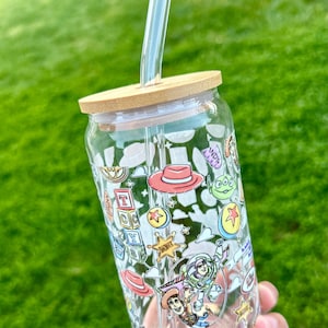 May include: A clear glass tumbler with a bamboo lid and a clear straw. The tumbler is decorated with colorful cartoon characters and icons. The design includes a red hat, a green alien, and the text "ANDY"