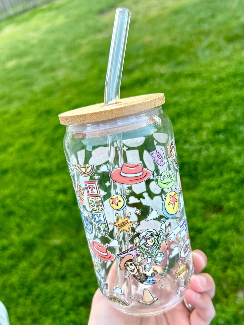 May include: A clear glass tumbler with a bamboo lid and a clear straw. The tumbler is decorated with colorful cartoon characters and icons. The design includes a red hat, a green alien, and the text "ANDY"