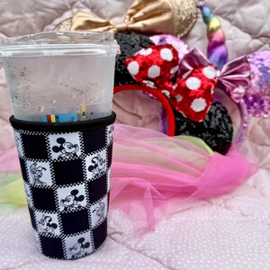May include: A clear plastic cup with a black and white Mickey Mouse checkered sleeve. Several character-themed headbands with bows and a unicorn horn are in the background, resting on a pink surface.