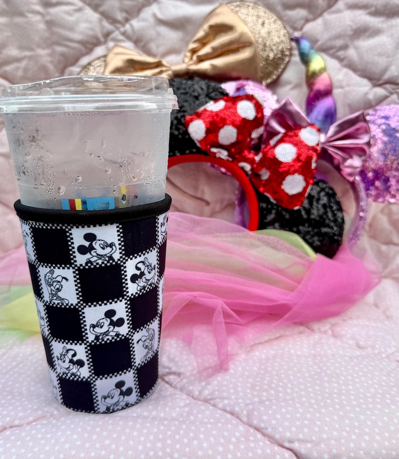 May include: A clear plastic cup with a black and white Mickey Mouse checkered sleeve. Several character-themed headbands with bows and a unicorn horn are in the background, resting on a pink surface.