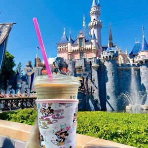 May include: A clear plastic cup with a pink straw, filled with a coffee drink, and a decorative sleeve featuring Mickey and Minnie Mouse characters. The background shows a fairytale castle under a bright blue sky.