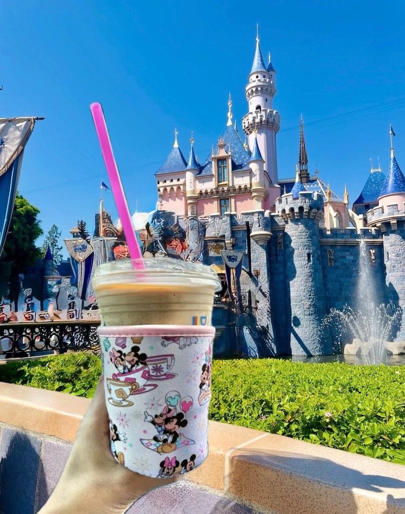 May include: A clear plastic cup with a pink straw, filled with a coffee drink, and a decorative sleeve featuring Mickey and Minnie Mouse characters. The background shows a fairytale castle under a bright blue sky.