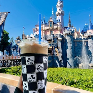 May include: A cold beverage in a clear plastic cup with a black and white Mickey Mouse checkered sleeve and a blue straw. The background features a castle with blue turrets and a fountain under a clear blue sky.