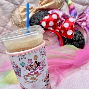 May include: A clear plastic cup with a blue straw, filled with a light brown beverage, sits in a pink and white patterned cup sleeve featuring Minnie and Mickey Mouse. Several character-themed headbands with bows are in the background.