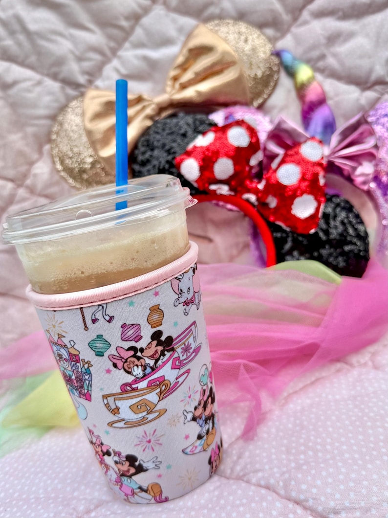 May include: A clear plastic cup with a blue straw, filled with a light brown beverage, sits in a pink and white patterned cup sleeve featuring Minnie and Mickey Mouse. Several character-themed headbands with bows are in the background.