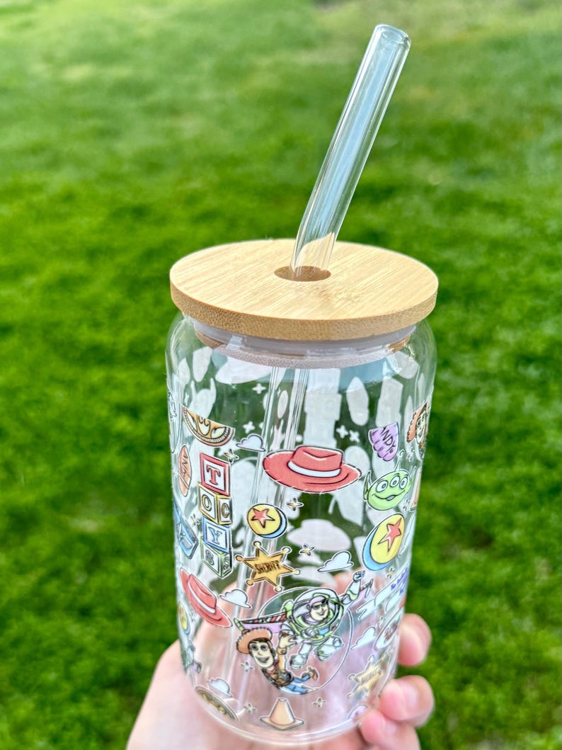 May include: A clear glass tumbler with a bamboo lid and a glass straw. The tumbler is decorated with colorful cartoon character graphics.