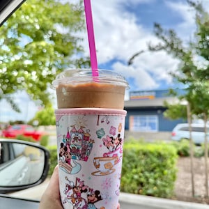 May include: A cold beverage in a clear plastic cup with a pink straw, held in a Disney-themed cup sleeve. The sleeve features cartoon characters and amusement park imagery in pink, blue, and white. The drink is brown, and the background shows trees and a blue sky.