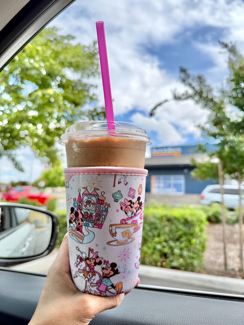 May include: A cold beverage in a clear plastic cup with a pink straw, held in a Disney-themed cup sleeve. The sleeve features cartoon characters and amusement park imagery in pink, blue, and white. The drink is brown, and the background shows trees and a blue sky.
