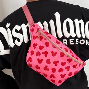 Disney Fanny pack with pink primary color with red hearts and Mickey Mouse symbol across Fanny pack in a scattered pattern. the strap is also pink and matches Fanny pack. it has a gold zipper.