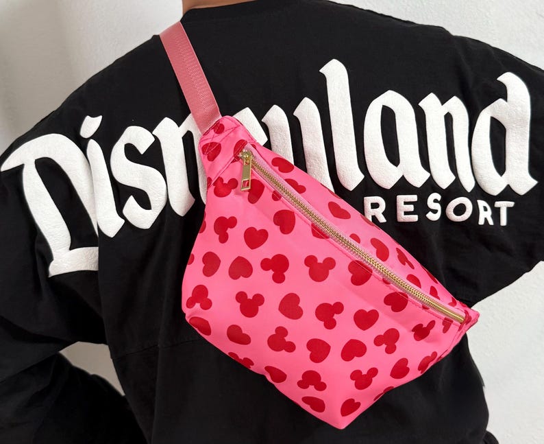 Disney Fanny pack with pink primary color with red hearts and Mickey Mouse symbol across Fanny pack in a scattered pattern. the strap is also pink and matches Fanny pack. it has a gold zipper.