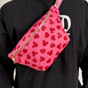Disney Fanny pack with pink primary color with red hearts and Mickey Mouse symbol across Fanny pack in a scattered pattern. the strap is also pink and matches Fanny pack. it has a gold zipper.