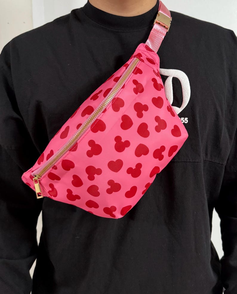 Disney Fanny pack with pink primary color with red hearts and Mickey Mouse symbol across Fanny pack in a scattered pattern. the strap is also pink and matches Fanny pack. it has a gold zipper.