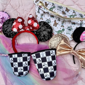 May include: A collection of themed headbands, including sequined ears, polka dot bows, and a gold glitter bow. Also included are two black and white checkered cups with Mickey Mouse stamps and a white fanny pack with a cartoon character print.