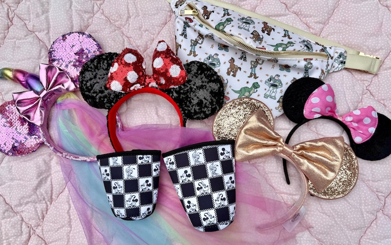 May include: A collection of themed headbands, including sequined ears, polka dot bows, and a gold glitter bow. Also included are two black and white checkered cups with Mickey Mouse stamps and a white fanny pack with a cartoon character print.