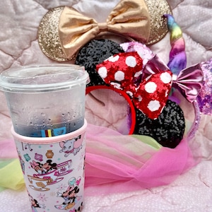 May include: A clear plastic cup with a beverage and a decorative sleeve featuring Disney characters. Several character-themed headbands with bows and ears are also visible, including a gold glitter bow and a red bow with white polka dots. The items are on a pink surface.