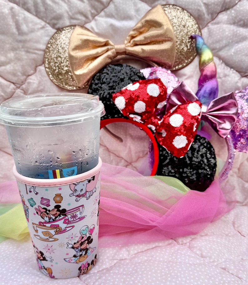 May include: A clear plastic cup with a beverage and a decorative sleeve featuring Disney characters. Several character-themed headbands with bows and ears are also visible, including a gold glitter bow and a red bow with white polka dots. The items are on a pink surface.