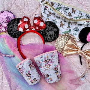 May include: A collection of themed headbands, including sequined unicorn ears, Minnie Mouse ears with red polka dots, and gold glitter ears. A white fanny pack with a Toy Story print and two patterned cups are also visible.