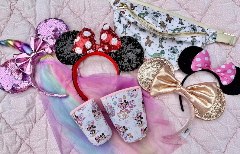 May include: A collection of themed headbands, including sequined unicorn ears, Minnie Mouse ears with red polka dots, and gold glitter ears. A white fanny pack with a Toy Story print and two patterned cups are also visible.