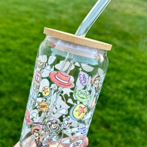 May include: A clear glass tumbler with a bamboo lid and a clear glass straw. The tumbler is decorated with colorful cartoon characters and phrases. The design includes a red cowboy hat, a green alien, and the words "To infinity and beyond.