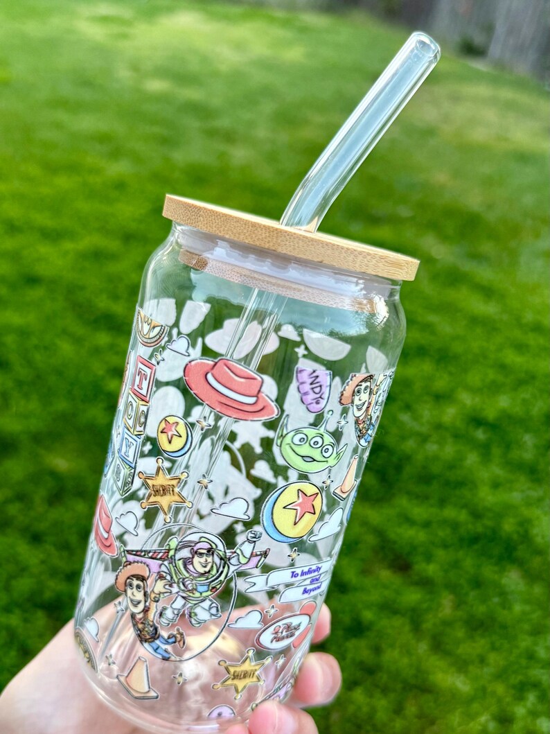 May include: A clear glass tumbler with a bamboo lid and a clear glass straw. The tumbler is decorated with colorful cartoon characters and phrases. The design includes a red cowboy hat, a green alien, and the words "To infinity and beyond.