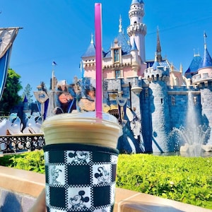 May include: A cold beverage in a clear plastic cup with a pink straw, held in a black and white Mickey Mouse checkered sleeve. The background features a fairytale castle with blue turrets and a fountain under a bright blue sky.