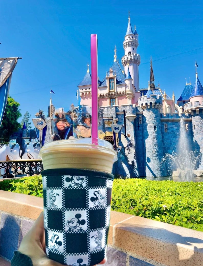 May include: A cold beverage in a clear plastic cup with a pink straw, held in a black and white Mickey Mouse checkered sleeve. The background features a fairytale castle with blue turrets and a fountain under a bright blue sky.