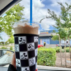 May include: A cold beverage in a clear plastic cup with a blue straw, held in a black and white checkered sleeve featuring Mickey Mouse and Minnie Mouse. The drink is a light brown color, and the background shows trees and a blue building.