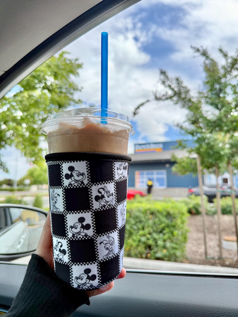 May include: A cold beverage in a clear plastic cup with a blue straw, held in a black and white checkered sleeve featuring Mickey Mouse and Minnie Mouse. The drink is a light brown color, and the background shows trees and a blue building.