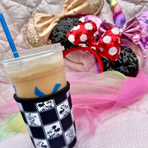 May include: A clear plastic cup with a blue straw, filled with a light brown beverage, sits in a black and white checkered sleeve featuring Mickey Mouse. Several character-themed headbands with bows and a unicorn horn are in the background.