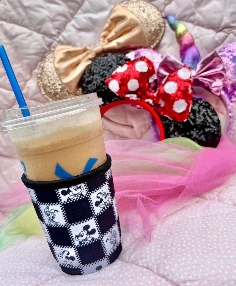 May include: A clear plastic cup with a blue straw, filled with a light brown beverage, sits in a black and white checkered sleeve featuring Mickey Mouse. Several character-themed headbands with bows and a unicorn horn are in the background.