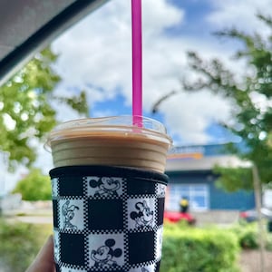 May include: Iced coffee in a clear plastic cup with a pink straw. The cup is in a black and white checkered sleeve featuring Mickey Mouse and other Disney characters. The background shows a blue sky with clouds and green trees.