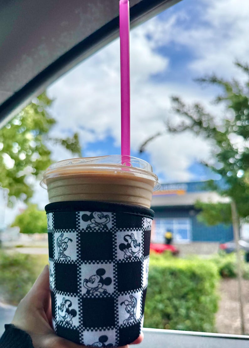 May include: Iced coffee in a clear plastic cup with a pink straw. The cup is in a black and white checkered sleeve featuring Mickey Mouse and other Disney characters. The background shows a blue sky with clouds and green trees.
