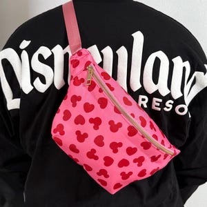 Disney Fanny pack with pink primary color with red hearts and Mickey Mouse symbol across Fanny pack in a scattered pattern. the strap is also pink and matches Fanny pack. it has a gold zipper.
