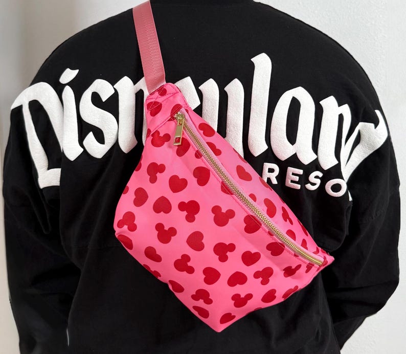 Disney Fanny pack with pink primary color with red hearts and Mickey Mouse symbol across Fanny pack in a scattered pattern. the strap is also pink and matches Fanny pack. it has a gold zipper.