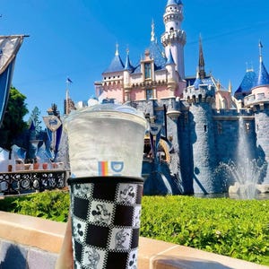 May include: A clear plastic cup with a lid and a black and white checkered sleeve featuring Mickey Mouse illustrations. The cup is held in front of a castle with blue turrets and a bright blue sky.
