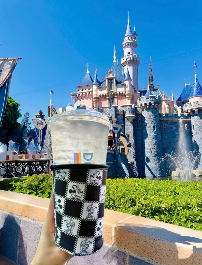 May include: A clear plastic cup with a lid and a black and white checkered sleeve featuring Mickey Mouse illustrations. The cup is held in front of a castle with blue turrets and a bright blue sky.