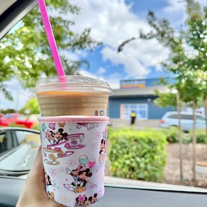 May include: A clear plastic cup filled with a coffee drink and a pink straw. The cup is in a white and pink sleeve featuring Mickey and Minnie Mouse illustrations. The background shows a sunny day with trees and a building.