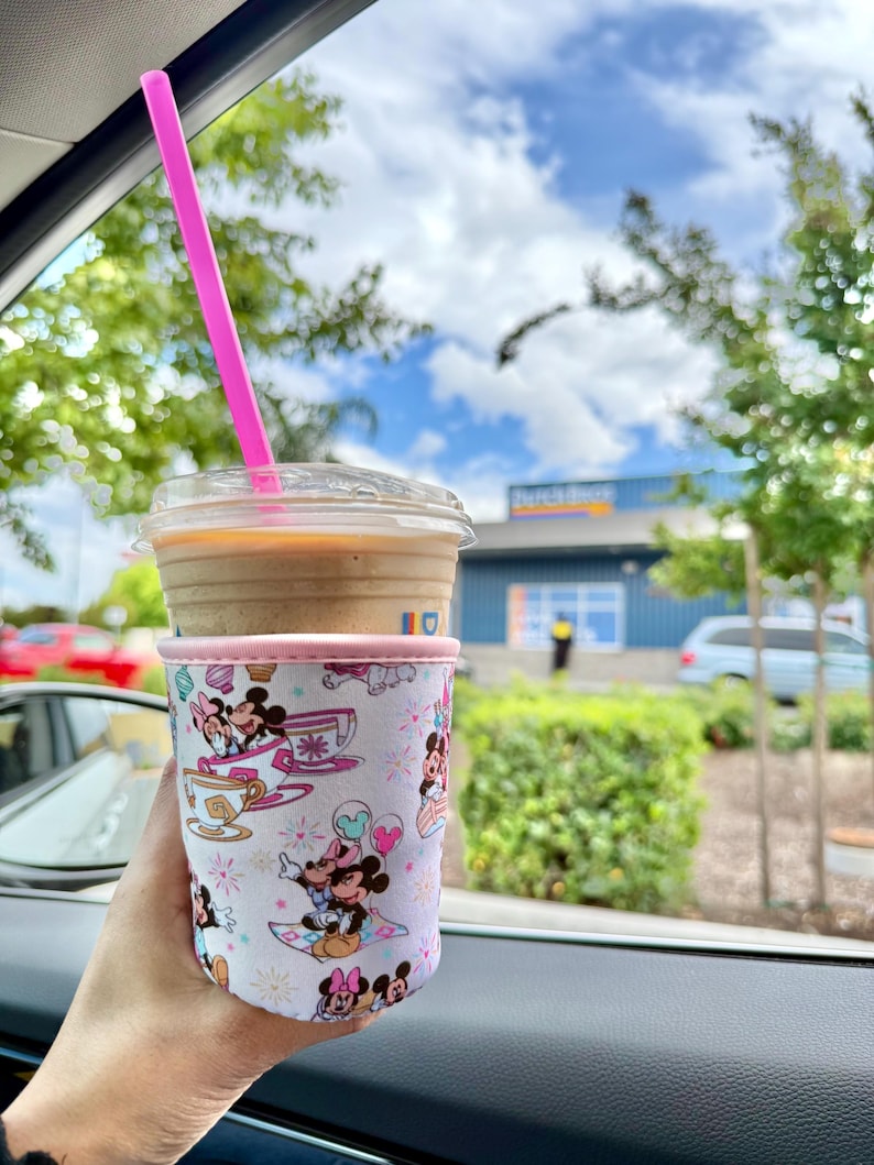 May include: A clear plastic cup filled with a coffee drink and a pink straw. The cup is in a white and pink sleeve featuring Mickey and Minnie Mouse illustrations. The background shows a sunny day with trees and a building.