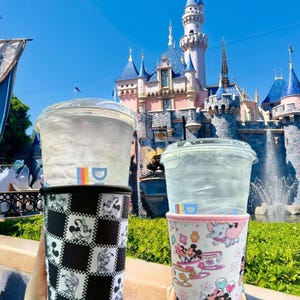 May include: Two beverage cup sleeves with Disney character designs. One sleeve features a black and white checkerboard pattern with Mickey Mouse illustrations. The other sleeve has a light pink background with various Disney characters.