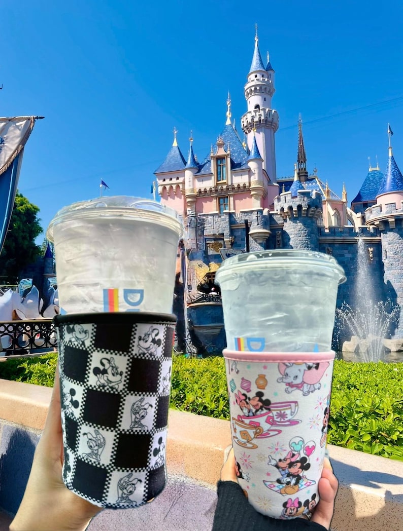 May include: Two beverage cup sleeves with Disney character designs. One sleeve features a black and white checkerboard pattern with Mickey Mouse illustrations. The other sleeve has a light pink background with various Disney characters.