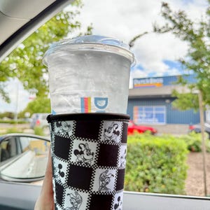 May include: A clear plastic cup filled with ice water, topped with a clear lid, and held in a black and white checkered sleeve featuring Mickey Mouse and friends. The cup is held in a car, with a view of a blue building and green trees.