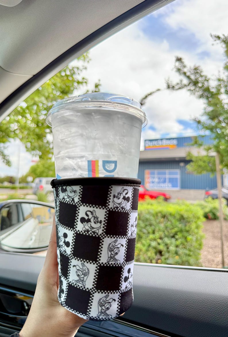 May include: A clear plastic cup filled with ice water, topped with a clear lid, and held in a black and white checkered sleeve featuring Mickey Mouse and friends. The cup is held in a car, with a view of a blue building and green trees.
