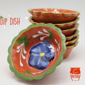 Puede incluir: Una pila de cuencos para salsas de color terracota con bordes festoneados y ribete verde. Cada cuenco presenta una flor azul pintada a mano con un centro amarillo. Las palabras "DIP DISH" están escritas en rojo.