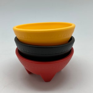 Puede incluir: Tres cuencos molcajete apilados, uno rojo, uno negro y uno amarillo. Los cuencos son de cerámica y tienen un diseño tradicional mexicano.