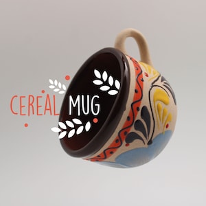 Puede incluir: Una taza de cerámica marrón y blanca con un diseño floral colorido. La taza está volcada, revelando el interior, que es marrón. El texto "CEREAL MUG" está escrito en letras blancas sobre un fondo rojo.