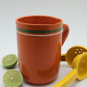 Puede incluir: Una taza de cerámica naranja con asa, con una franja verde y blanca cerca del borde. Dos mitades de lima y dos exprimidores de cítricos amarillos también están en la imagen. La taza es probablemente hecha a mano.