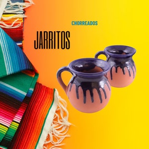 Puede incluir: Dos jarritos tradicionales mexicanos de barro, con un esmalte morado y un interior rosa. Los jarritos están sobre un fondo amarillo con un sarape colorido en la esquina. El texto "CHORRREADOS" y "JARRITOS" es visible en la imagen.
